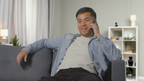Young-Man-Sitting-On-Sofa-At-Home-Smiling-And-Talking-With-Friend-On-Mobile-Phone